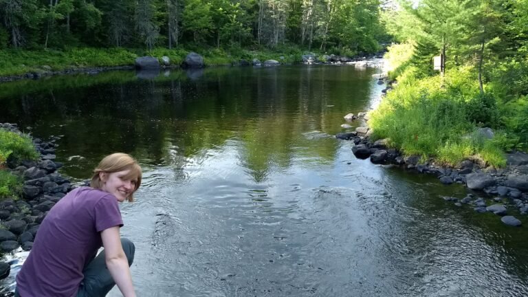 A woman sits on a rook overlooking a pool of the Pleasant River