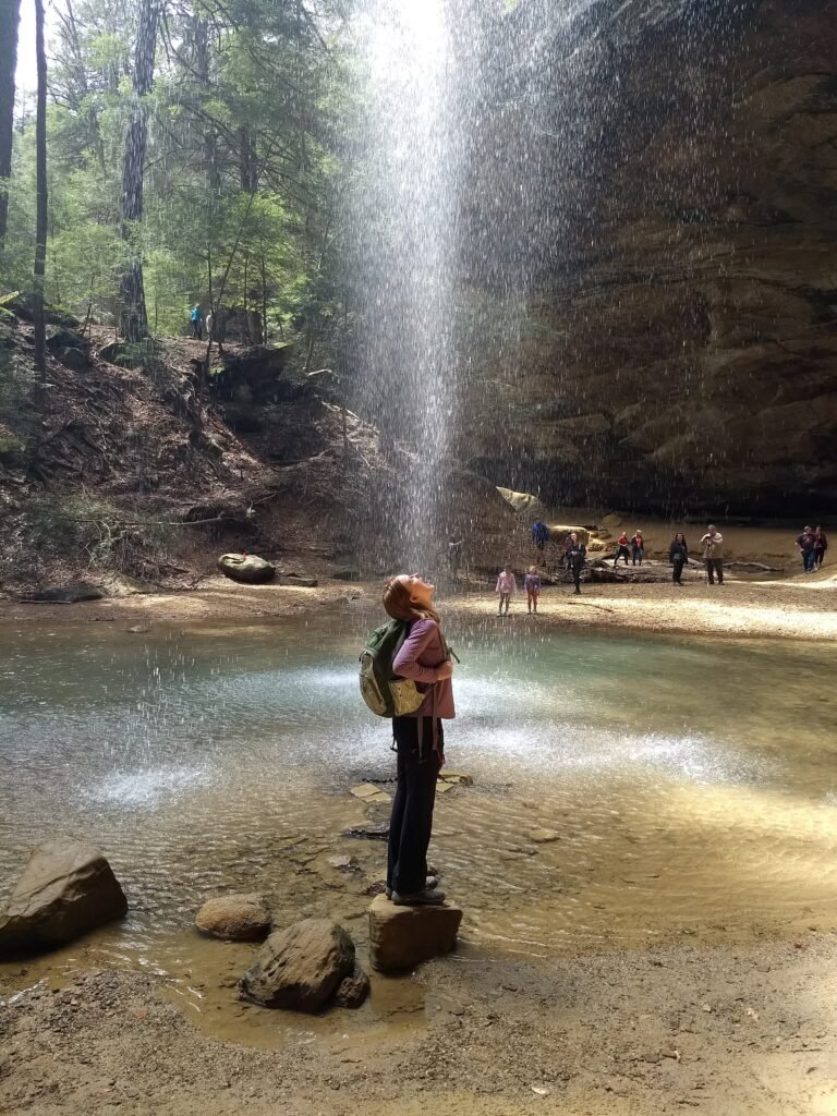 A woman stands on a rock under the lip of a massive recessed cave, as a shower of water falls from the top. Sunlight from behind the water illuminates the shower, as if it's a beam of energy